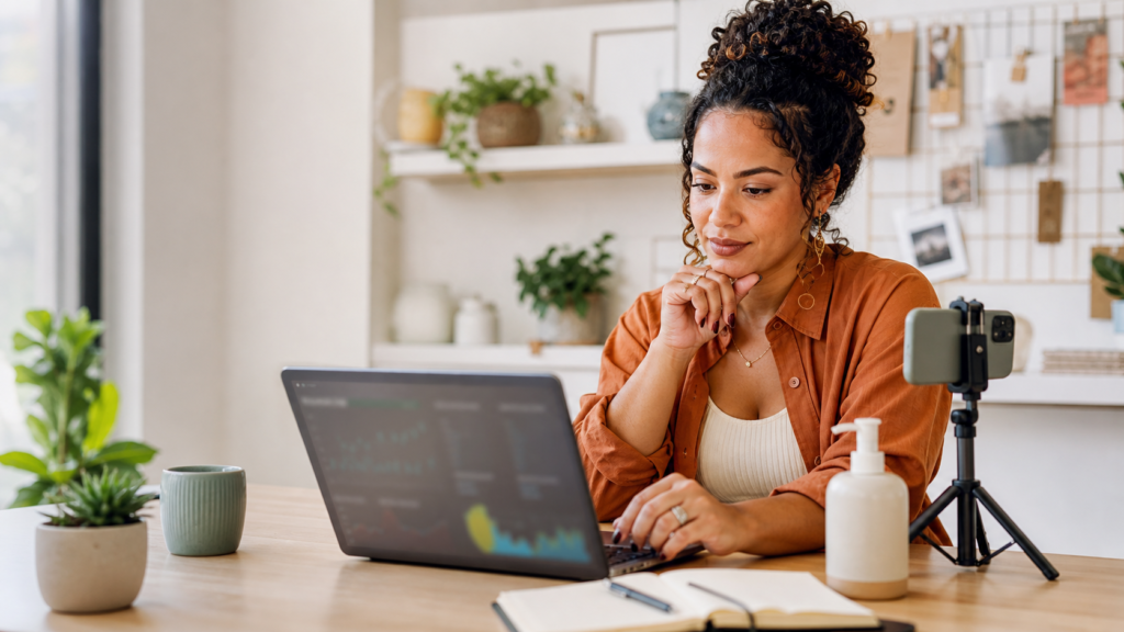 Criadora analisando o desempenho de recomendações de produtos em um laptop, com celular em tripé ao lado.